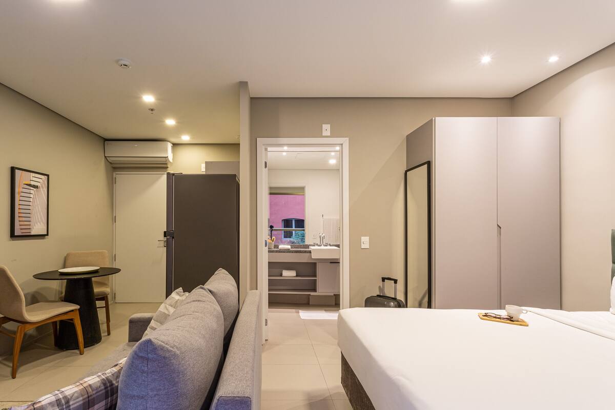 Ein modernes Hotelzimmer mit einem Bett, einer Sitzecke mit Tisch und Stühlen, einem Kleiderschrank und einem durch eine offene Tür einsehbaren Badezimmer.