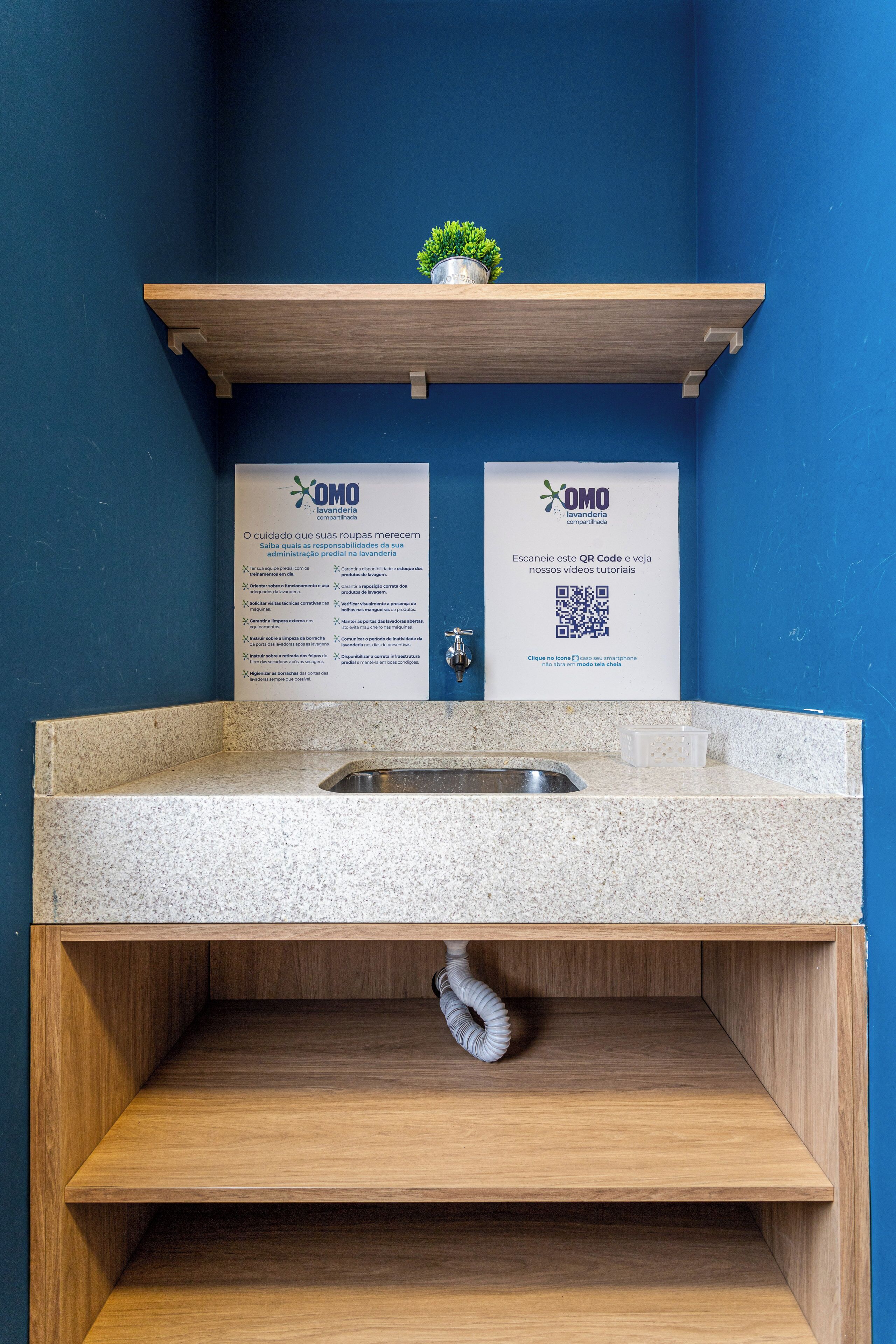 A bathroom with a blue wall, a wooden shelf with a potted plant, a sink with a granite countertop, and two informational posters with QR codes.