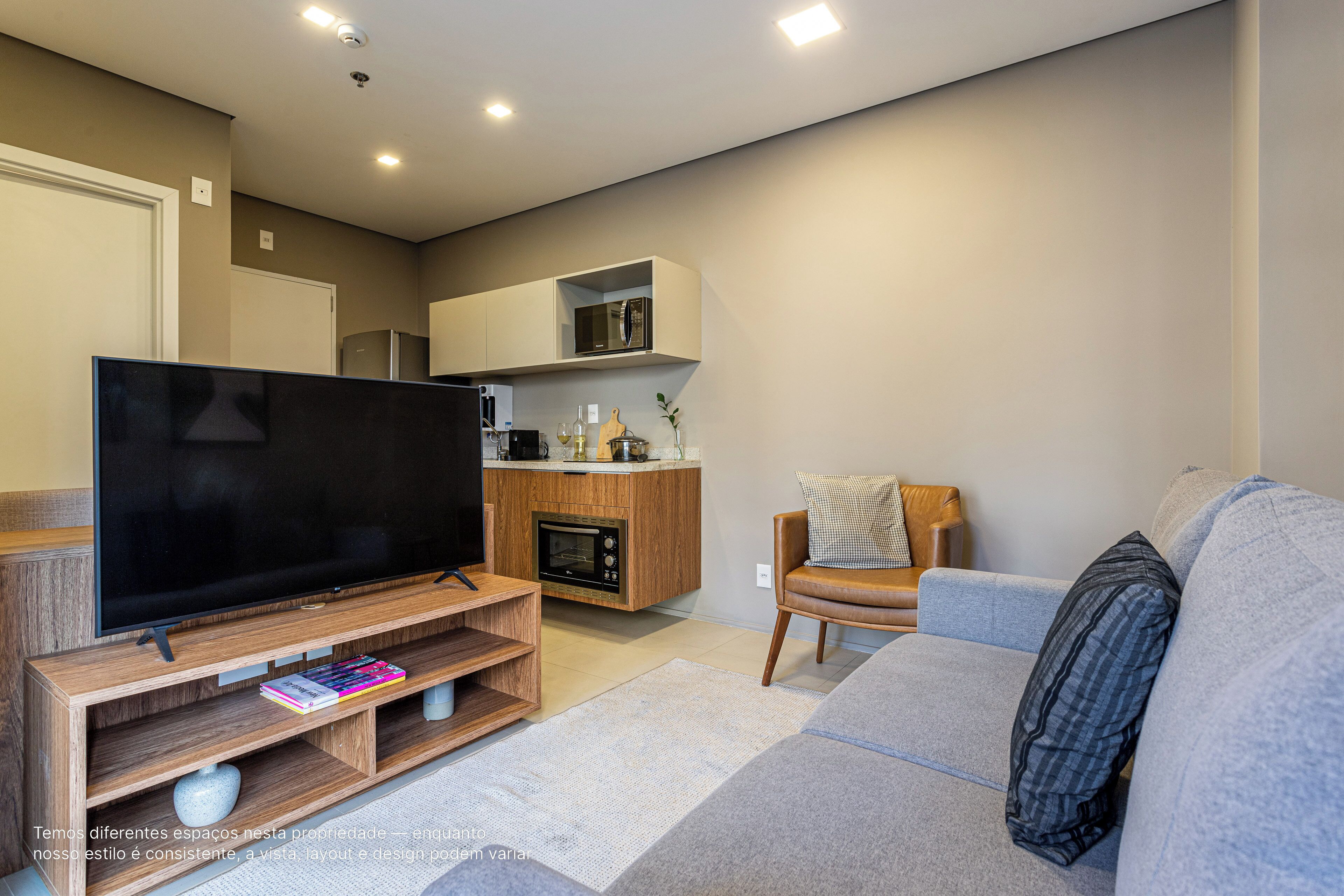 A modern living room with a flat-screen TV, wooden entertainment unit, grey sofa, and a kitchen area with built-in appliances.