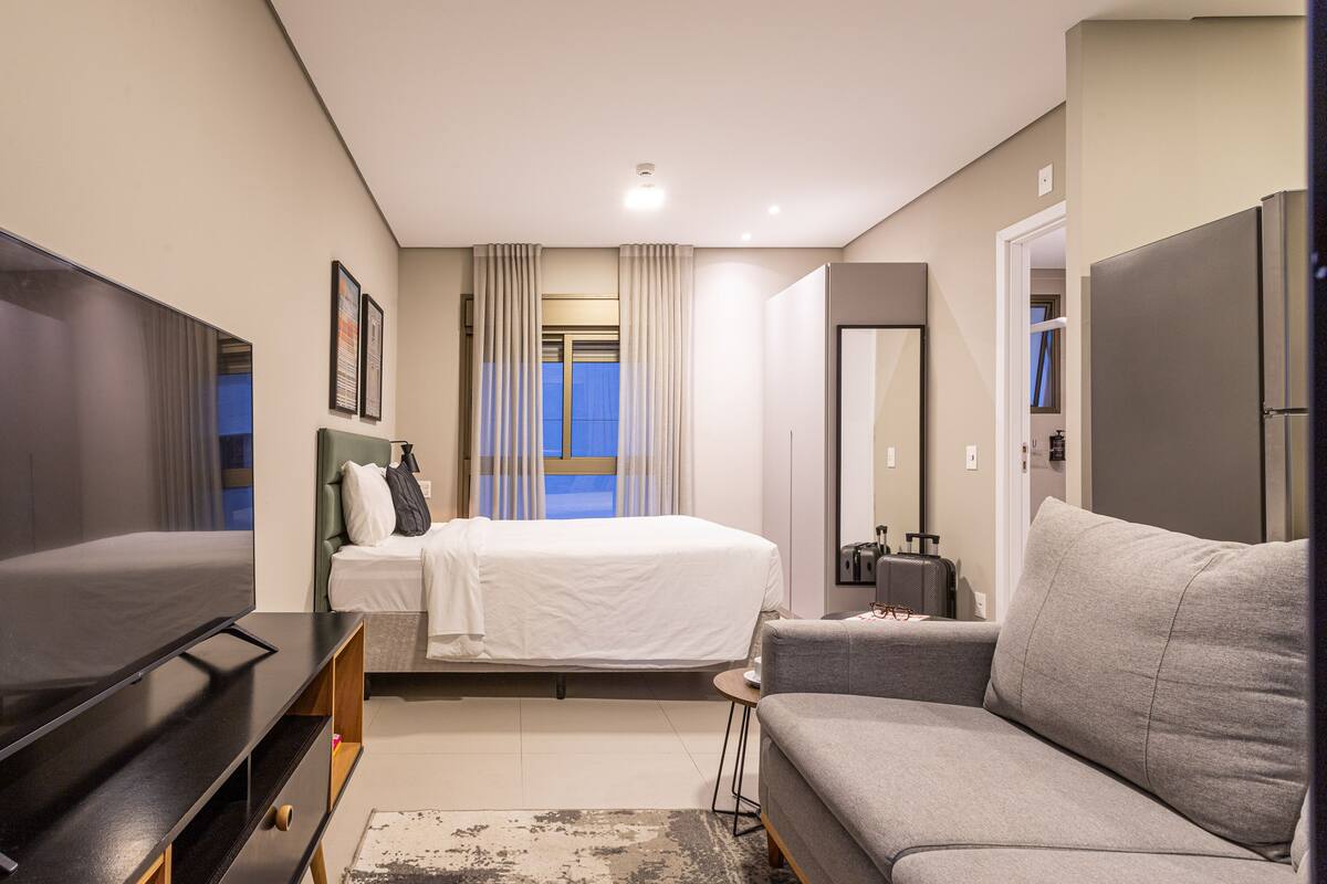 Ein modernes Hotelzimmer mit einem großen Flachbildfernseher, einem Bett mit weißer Bettwäsche, einem grauen Sofa und einem kleinen Beistelltisch.