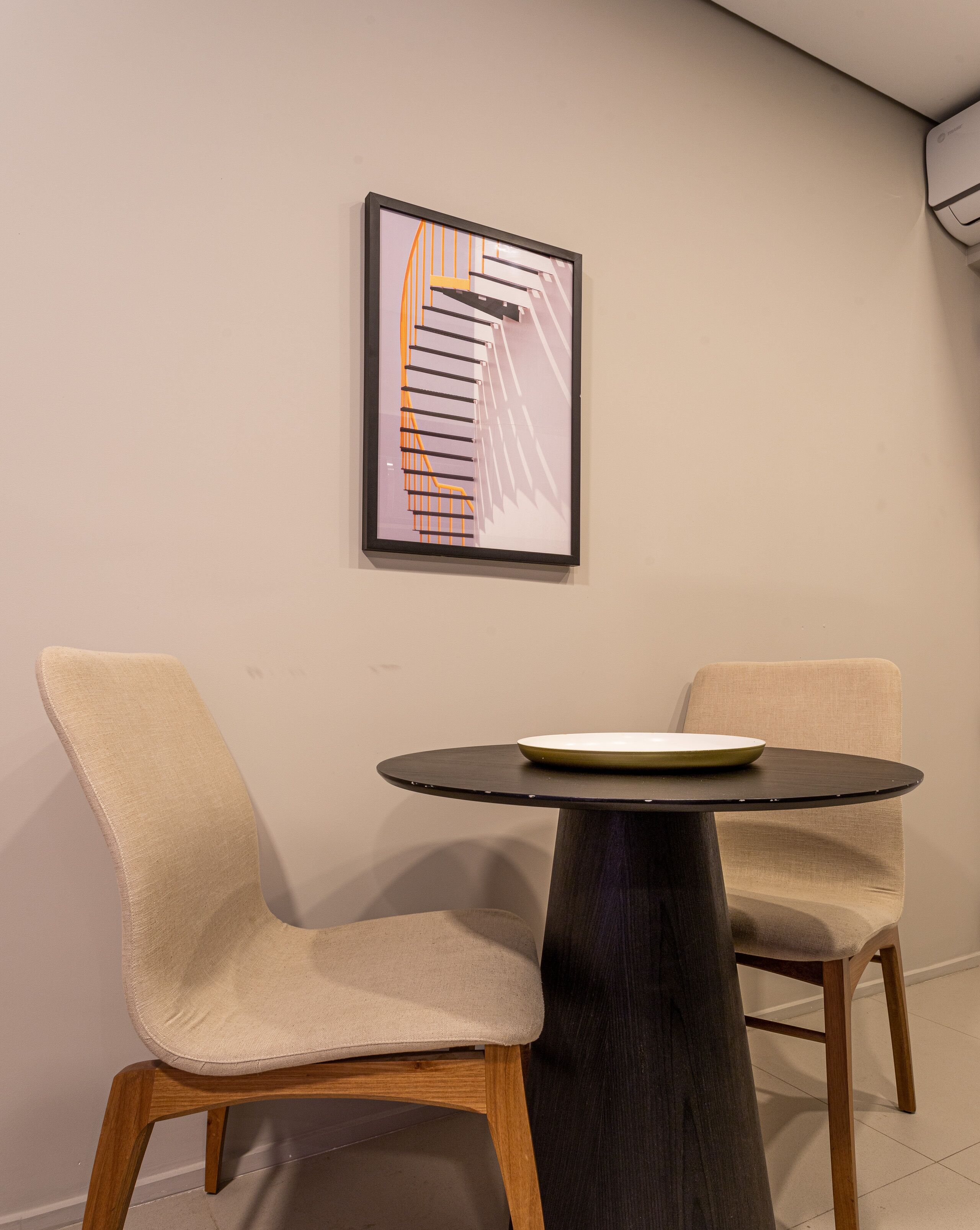 A modern room with a round table and two beige chairs. A framed abstract artwork is on the wall.
