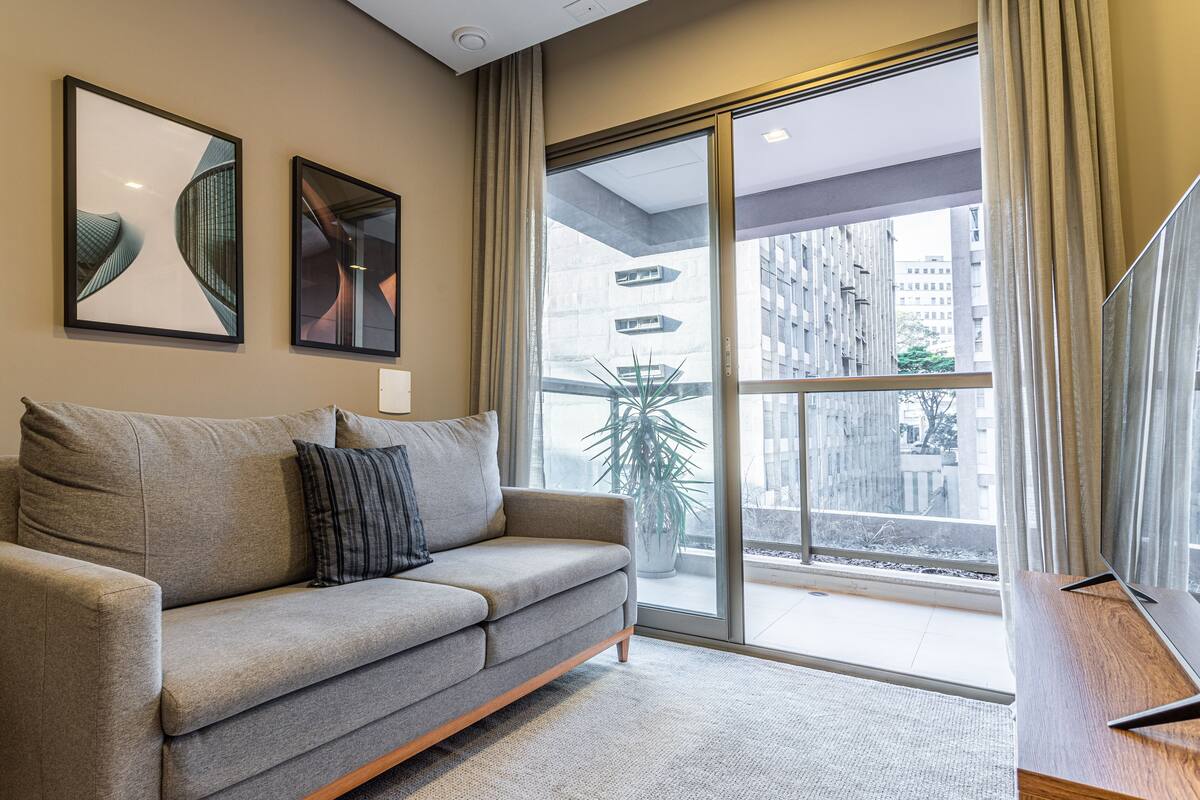 Ein modernes Wohnzimmer mit einem grauen Sofa, zwei gerahmten Kunstwerken, einem großen Fenster mit Blick auf Gebäude und einem Flachbildfernseher auf einem Holzboden.