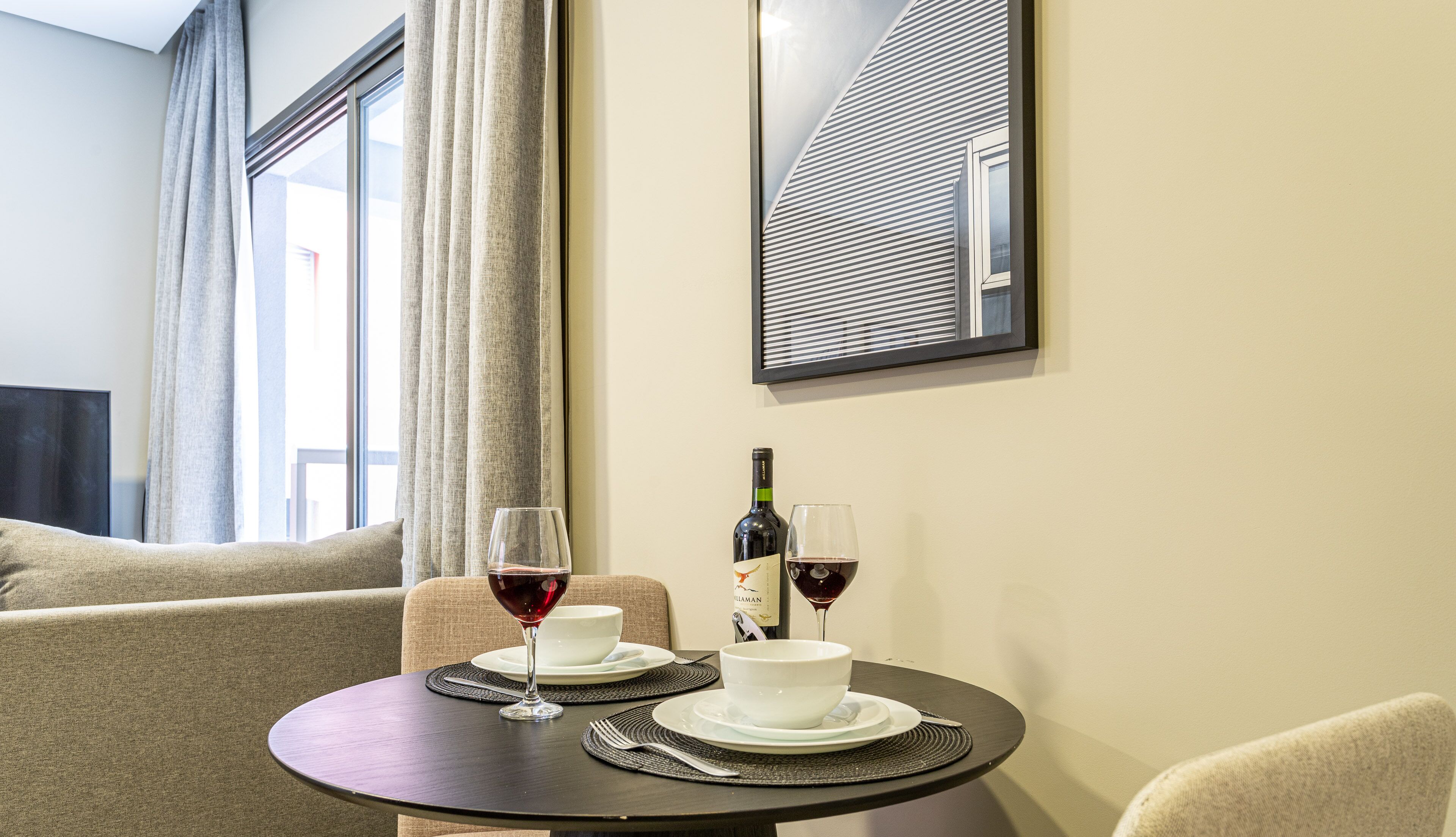 Un coin repas avec une table dressée pour deux personnes, deux verres à vin, une bouteille de vin et un tableau encadré accroché au mur.