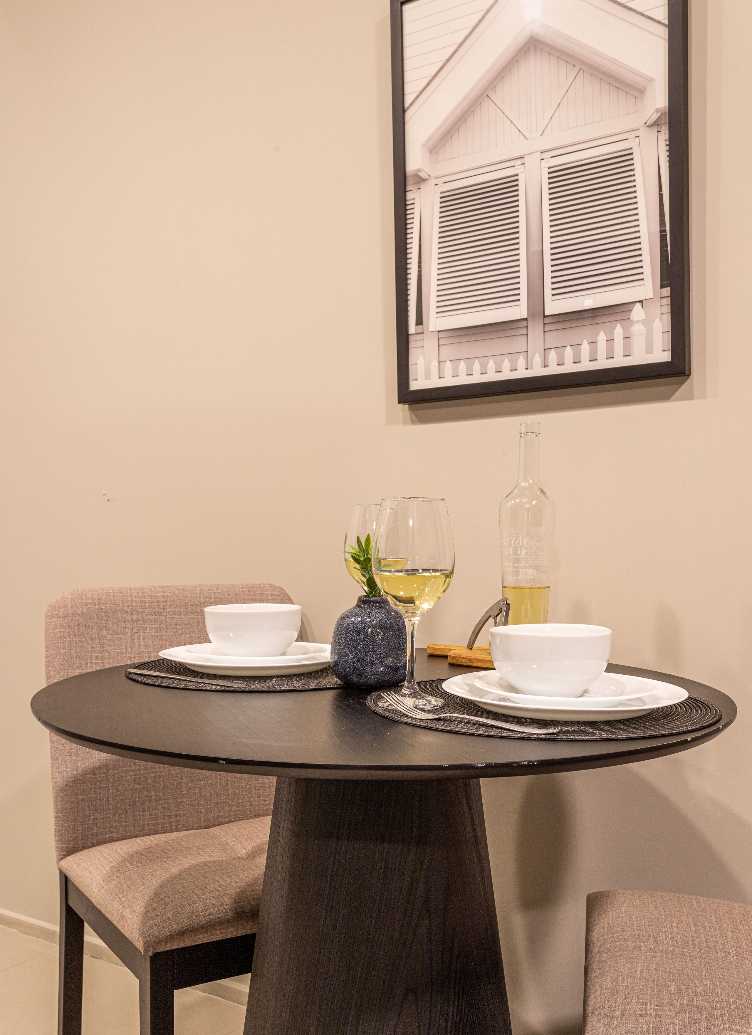 A round table with a dark wood finish, two chairs with light brown fabric, and a framed picture of a house with shutters on the wall behind.