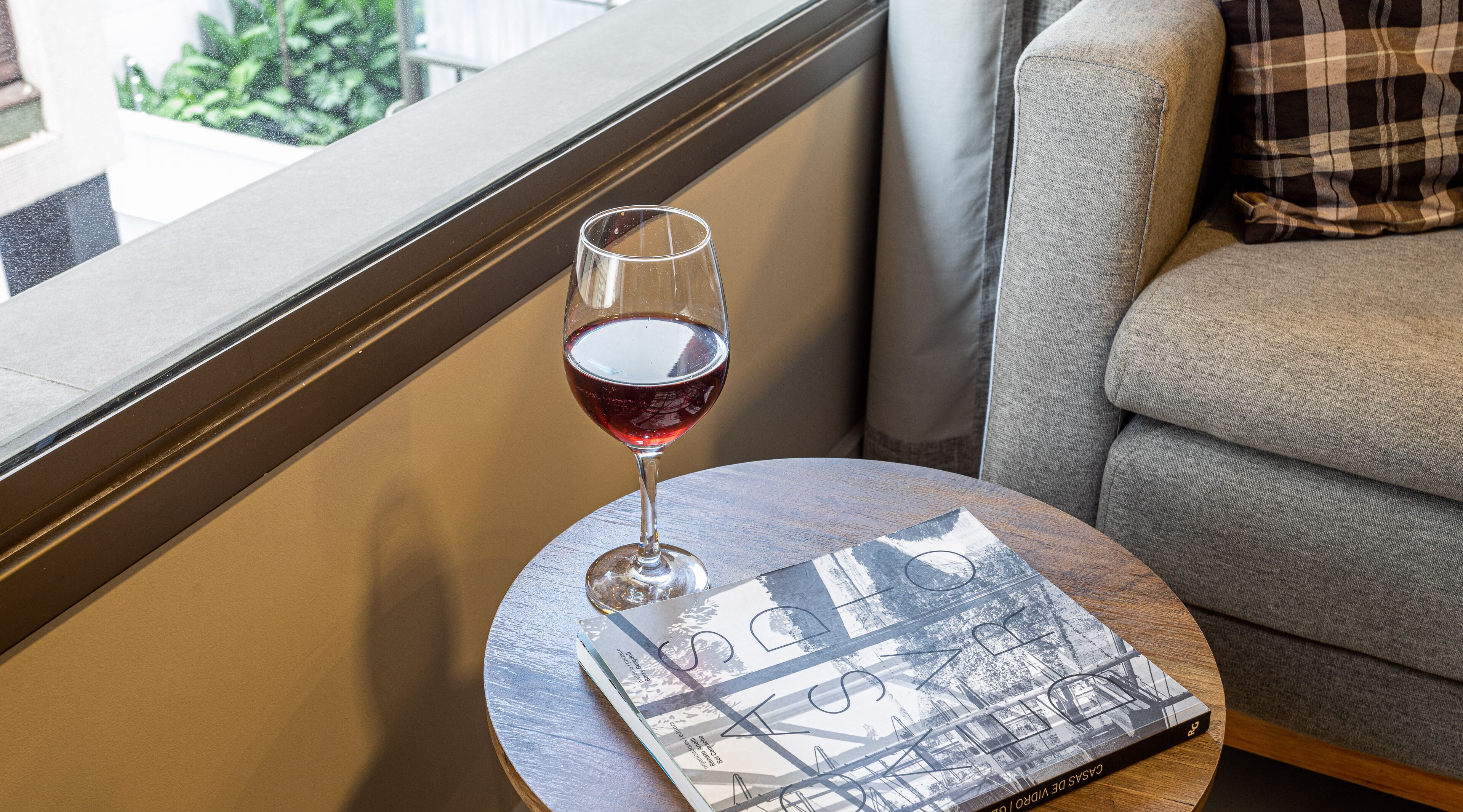 Una copa de vino tinto sobre una mesa redonda junto a un sofá con un cojín escocés.