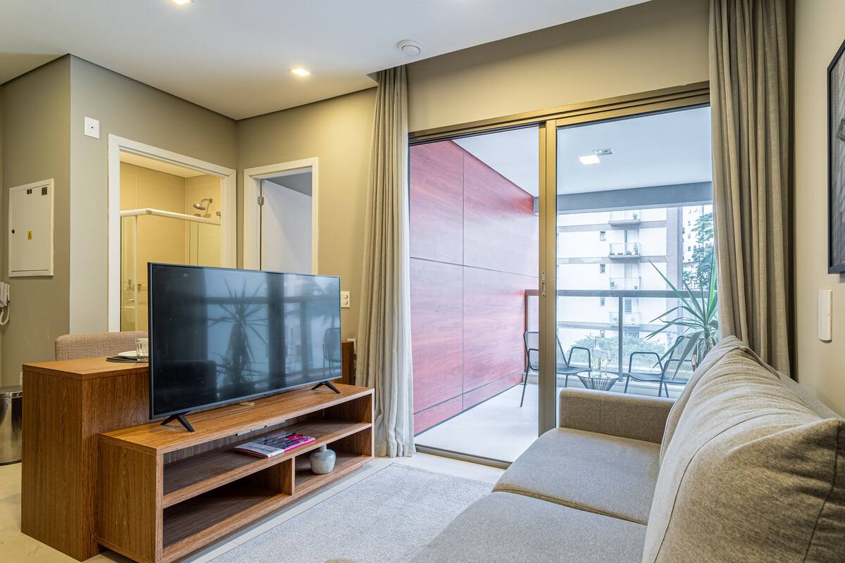 Ein modernes Wohnzimmer mit einem Flachbildfernseher auf einem Holzbodenständer, einer Couch und einer Glasschiebetür, die zu einem Außenbalkon führt.