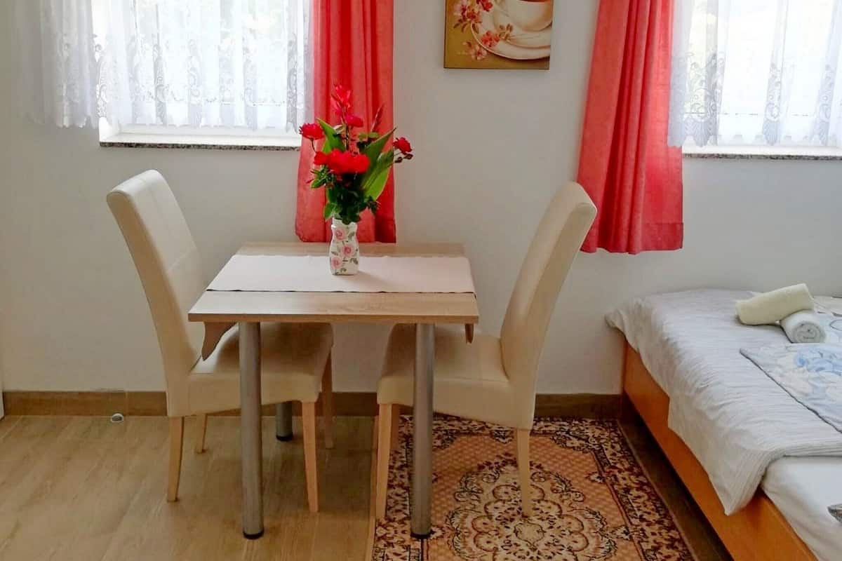 A room with a dining table, two chairs, a vase with flowers, a bed with a rolled-up towel, and a painting on the wall.