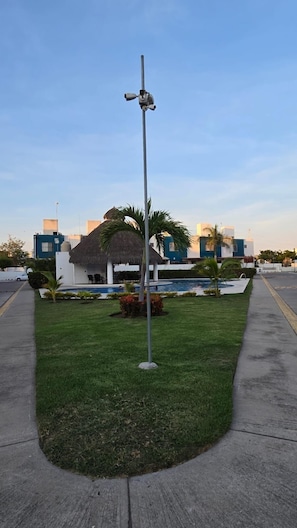 A grassy area with a pole, a pool, and modern buildings in the background.