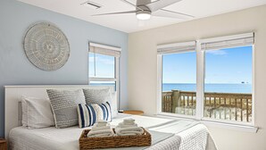 Une chambre à coucher avec un lit, des oreillers, un ventilateur de plafond et une vue sur la plage, visible à travers de grandes fenêtres.