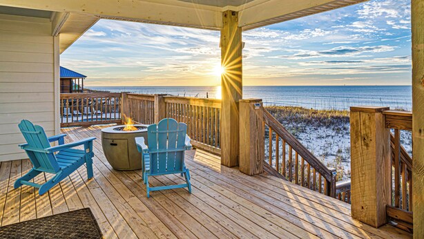 Une terrasse en bois avec deux fauteuils Adirondack et un foyer, offrant une vue sur une plage et l’océan.