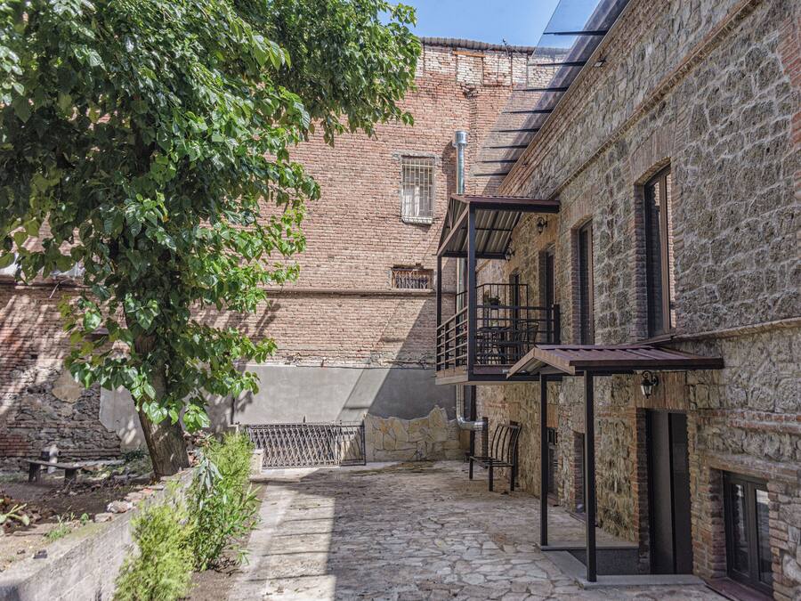 A courtyard with a cobblestone path, stone walls, and a tree providing shade.