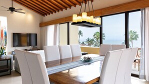 Un espace de restauration moderne avec une table en bois, des chaises à revêtement blanc et une vue sur l’océan grâce à de grandes fenêtres.