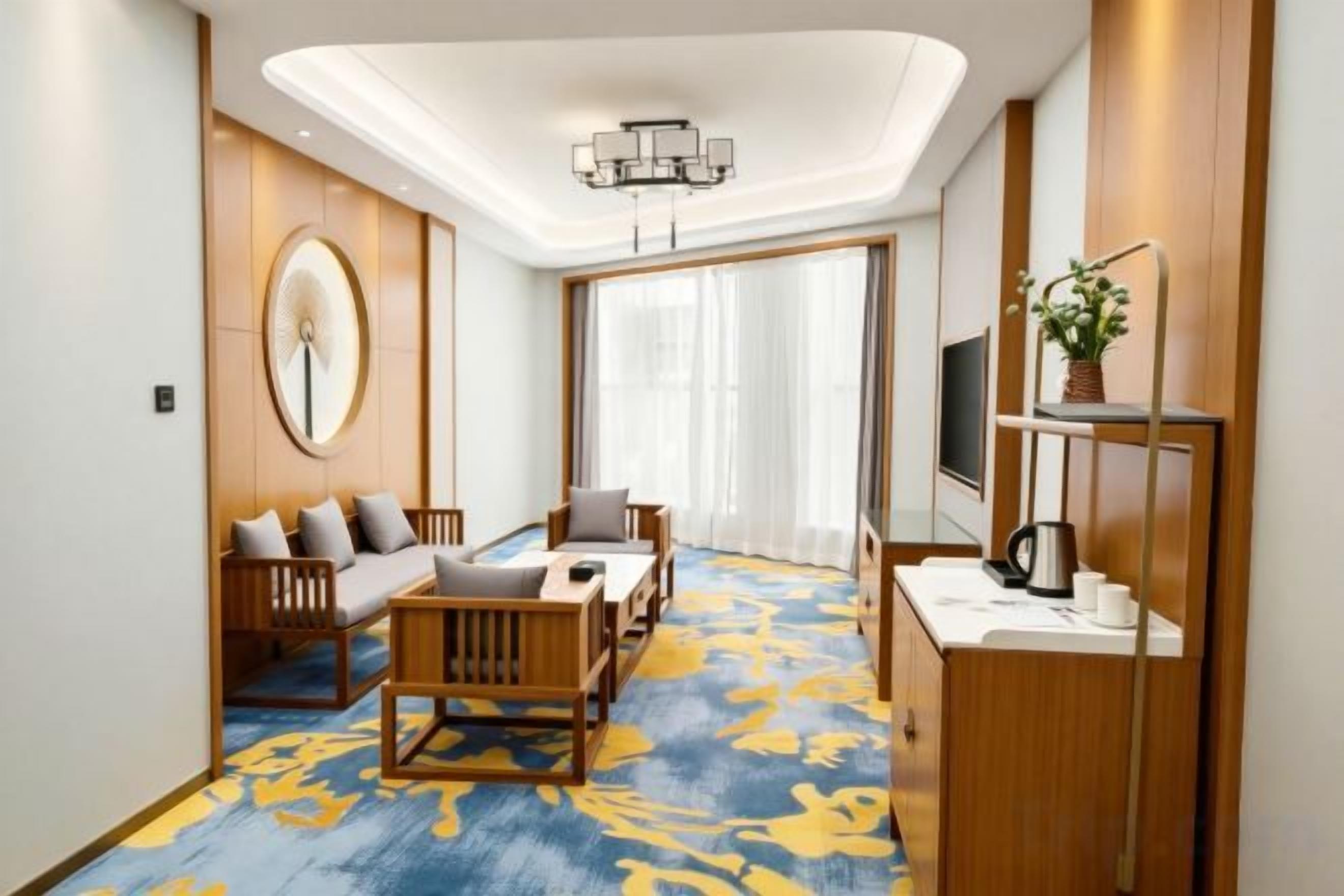 Ein Hotelzimmer mit einer Sofagarnitur, einem Couchtisch, einem Fernseher, einem Schreibtisch mit Computer und einem großen Fenster mit Vorhängen.