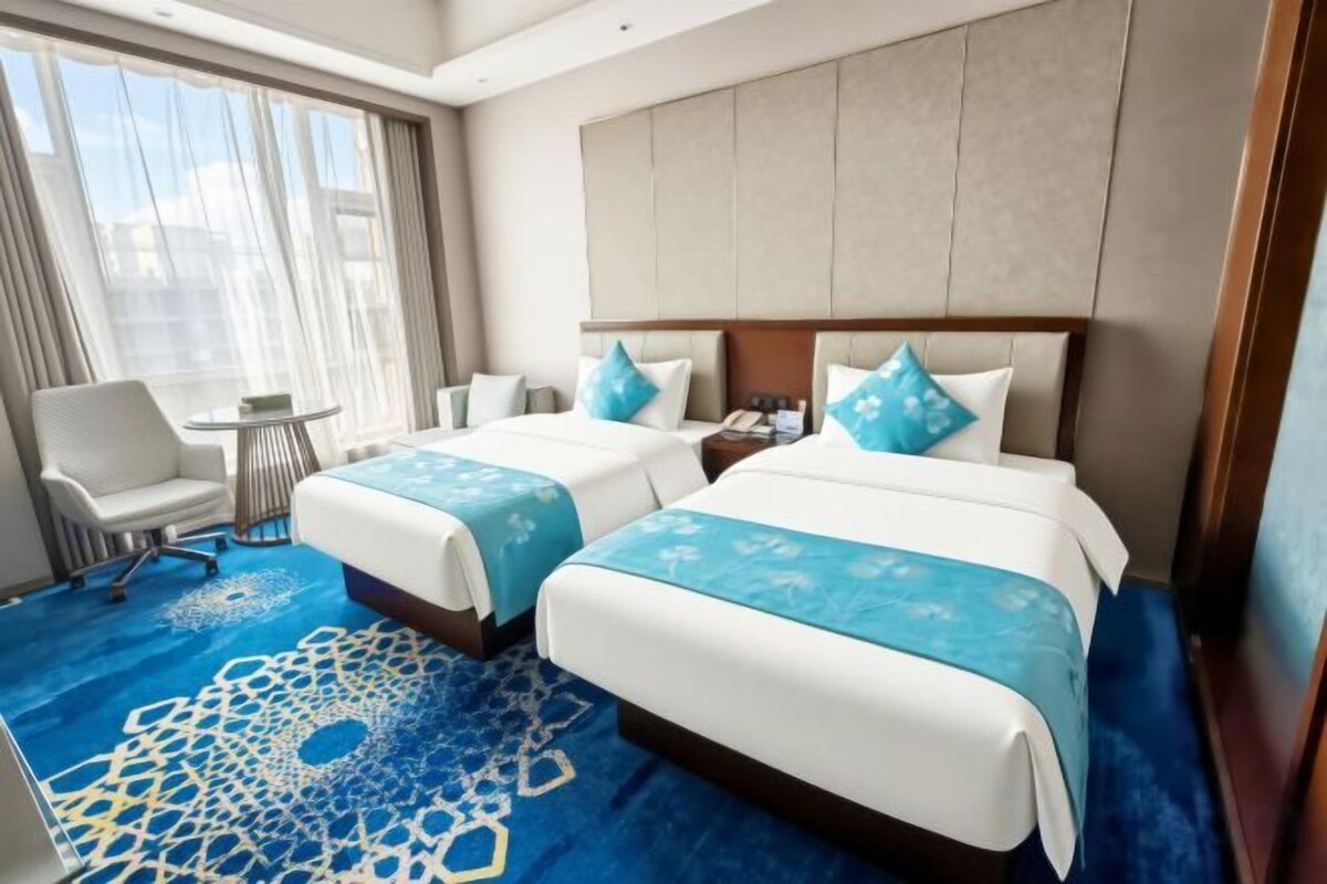 A hotel room with two beds, a desk, a chair, and a view of the city.