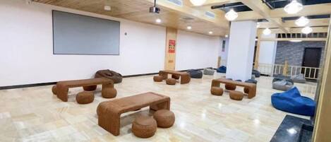 Una habitación amplia con techo de madera, pantalla de proyector y varias zonas de estar, incluyendo sillones de mimbre y un banco.