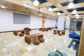 Una habitación amplia con techo de madera, pantalla de proyector y varias zonas de estar, incluyendo sillones de mimbre y un banco.
