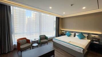A hotel room with a large bed, a small table, two armchairs, and a view of the city through large windows.