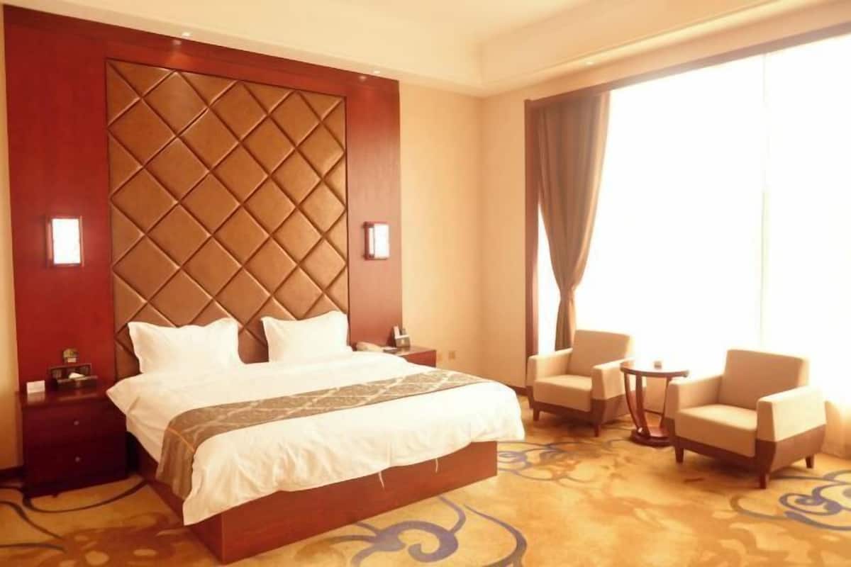 A hotel room with a large bed, two bedside tables, a small armchair, a round table, and a large window with curtains.
