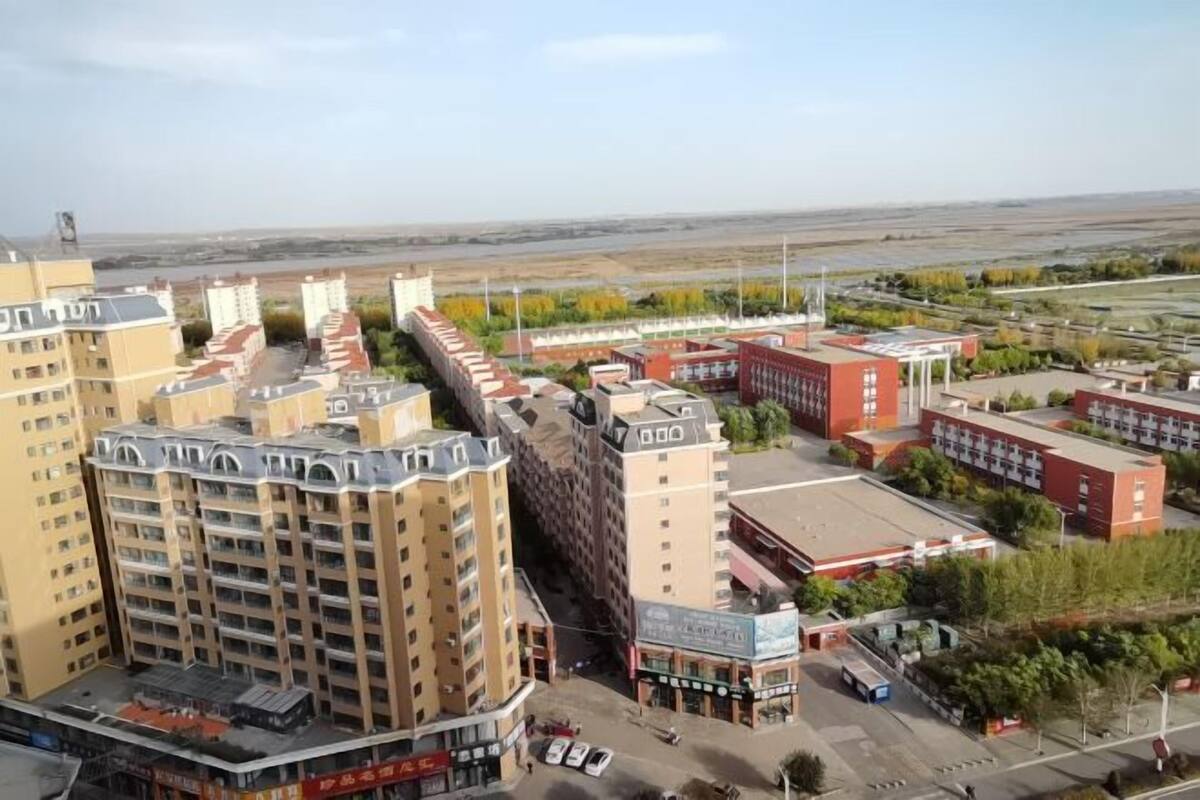Un edificio de varios pisos con techo plano, rodeado de otras construcciones y un terreno llano.