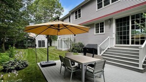 Una terraza con mesa y sillas, un paraguas amarillo y una casa con tejado rojo al fondo.