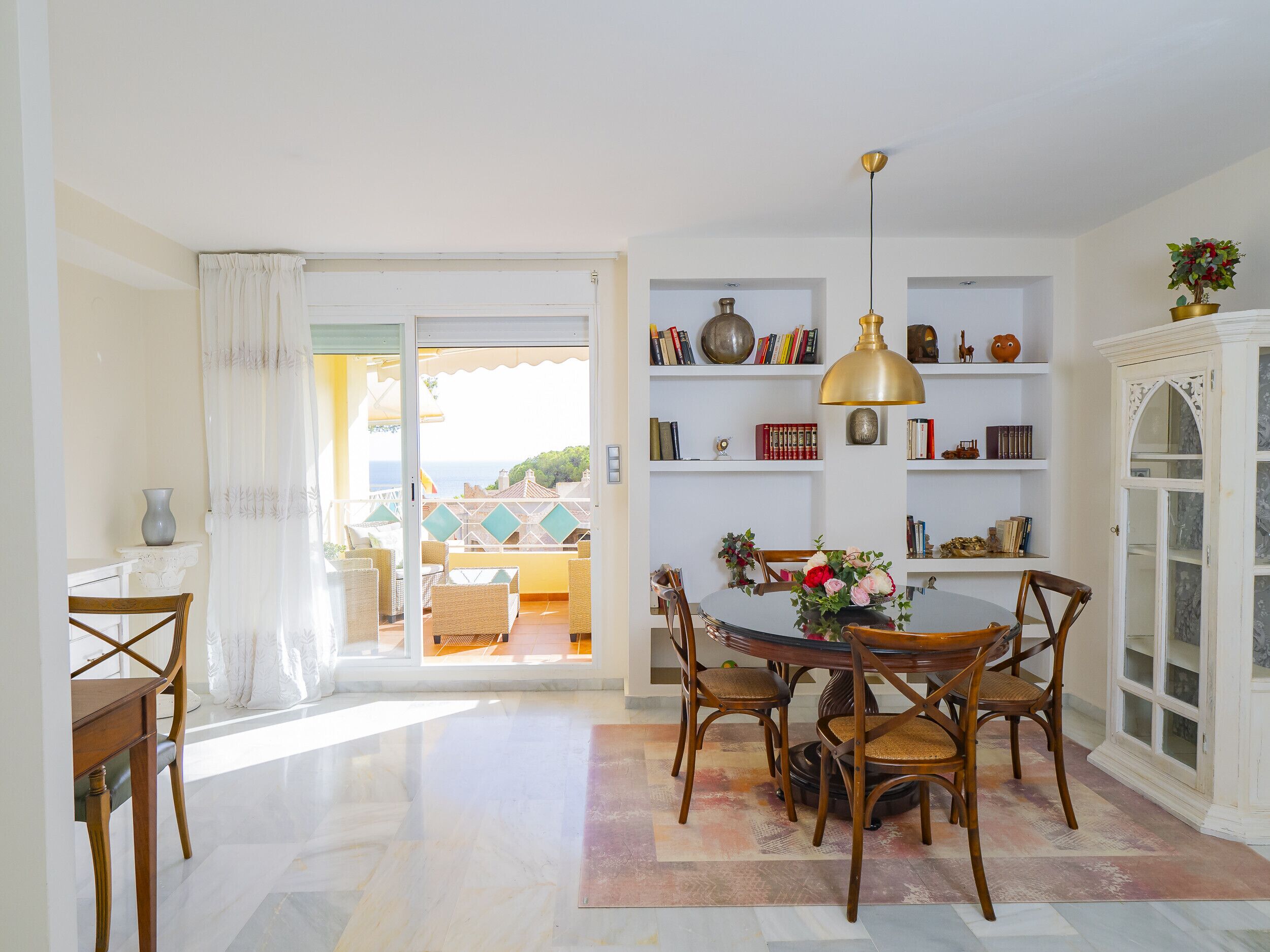 A dining area with a round table, chairs, and a chandelier. There is a bookshelf with books and decorative items. A sliding door leads to an outdoor area with a view of the sea.