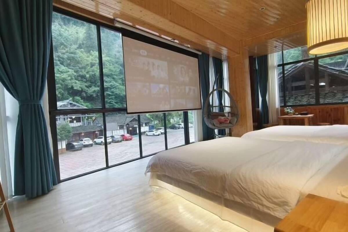 A room with a large bed, a projector screen, and a view of a parking area through large windows.