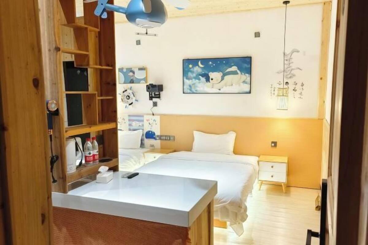 A hotel room with a bed, bedside table, nightstand, and a desk with a lamp. There is a painting on the wall and a shelf with various items.