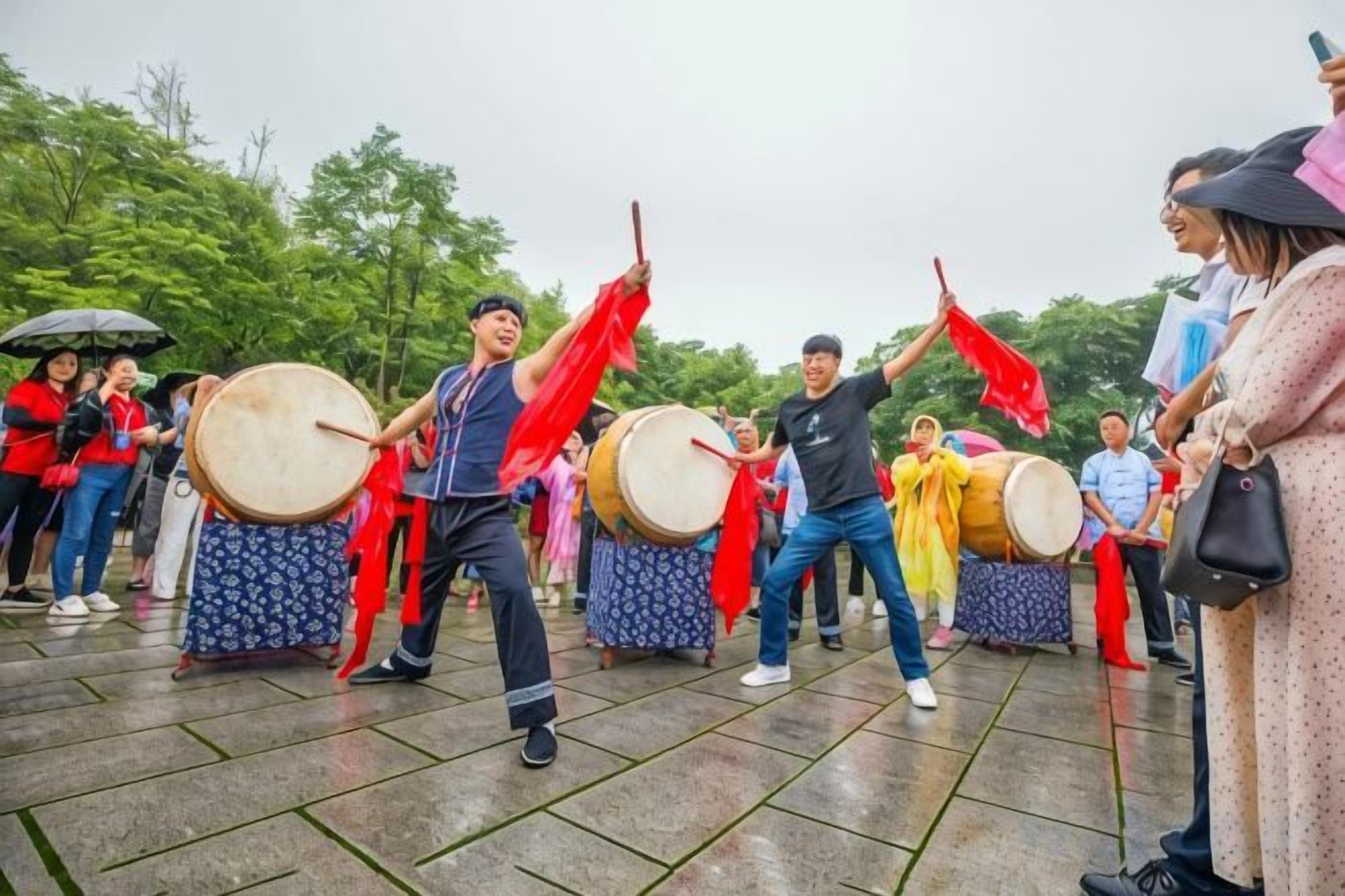 A group of people playing drums outdoors, with onlookers in the background.