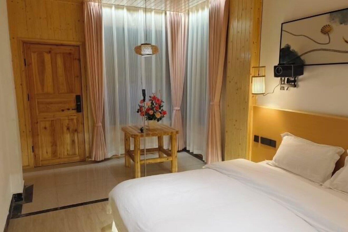 Habitación de hotel con puerta de madera, cama con ropa blanca, mesita de madera con un florero y una lámpara de pared.