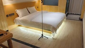 Una cama bien hecha con edredón blanco y almohadas, una mesita de noche de madera y una lámpara de pie.