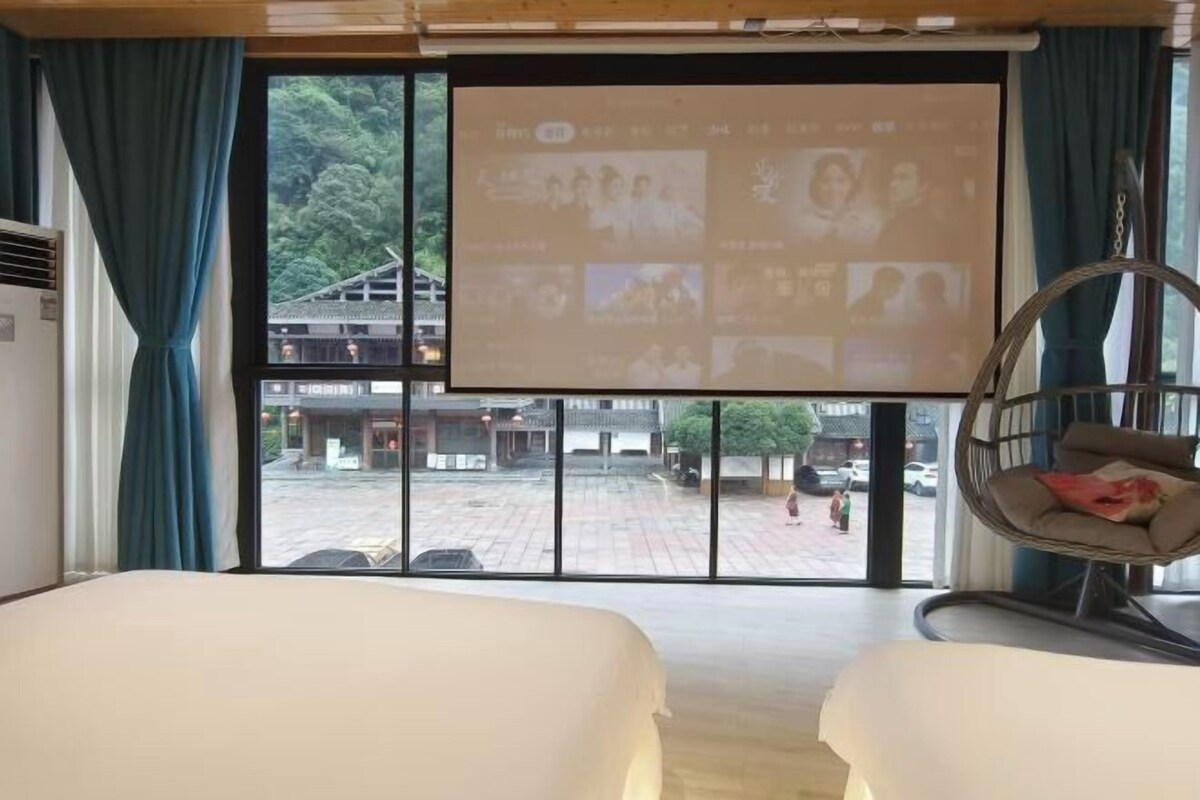 Habitación de hotel con un ventanal grande, una cama, una silla colgante y una pantalla de proyección mostrando imágenes.