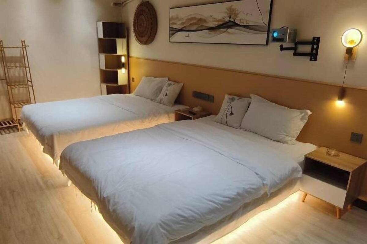 A hotel room with two beds, a bedside table, a wall-mounted shelf, and a wall-mounted light fixture.