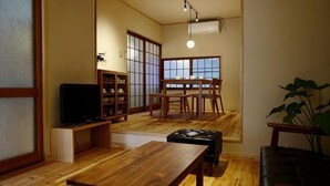Ein traditionelles japanisches Wohnzimmer mit Tatami-Böden, einem niedrigen Holztisch, einer schwarzen Ledersofa, einem Fernseher und einem Essbereich mit einem niedrigen Tisch und Stühlen.