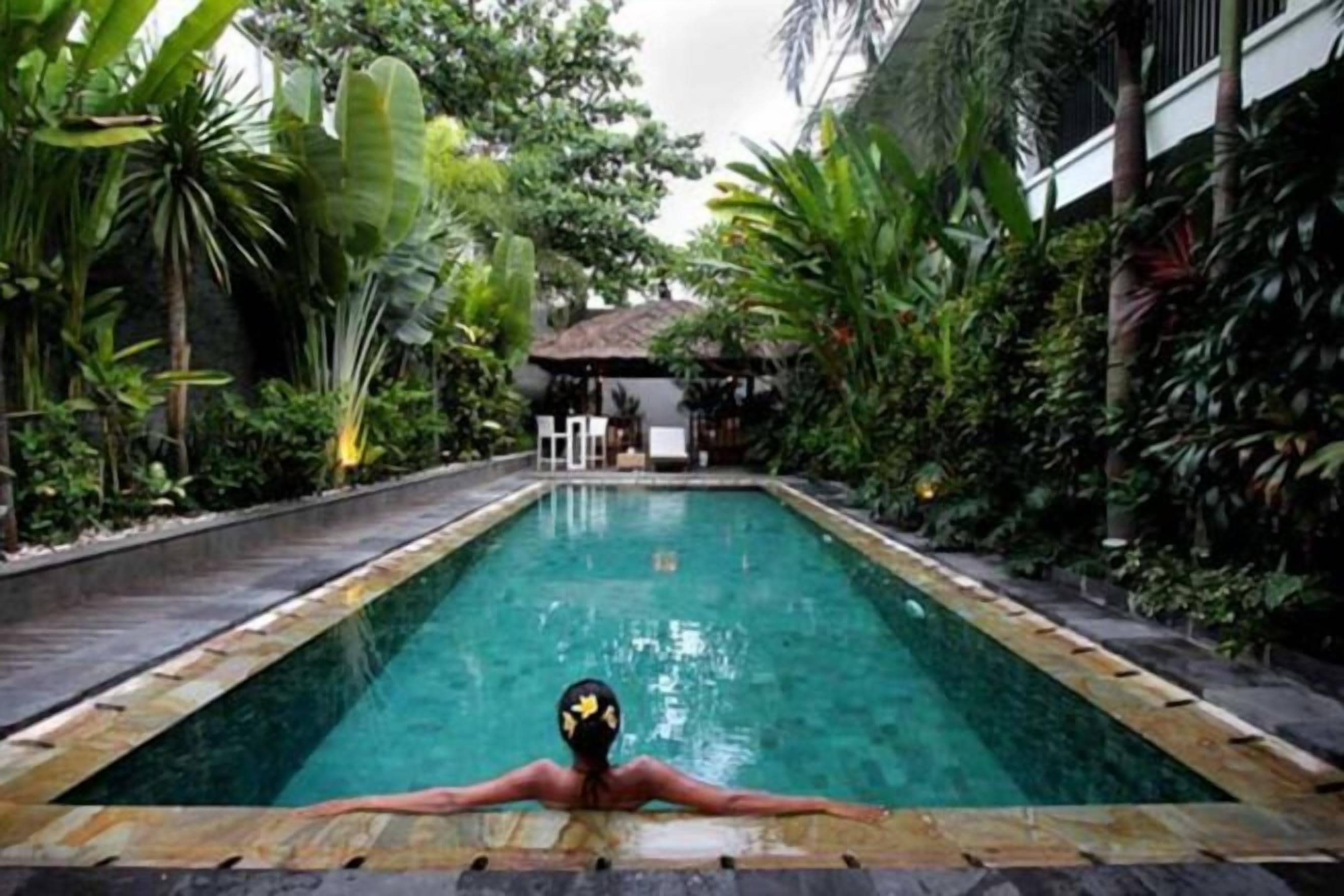 A rectangular pool with a person relaxing at the far end, surrounded by lush greenery and a building in the background.