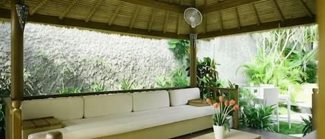 A covered outdoor seating area with a sofa and a table, surrounded by plants and a stone wall.