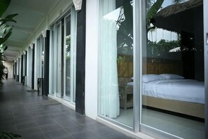 Un corridoio con un letto in una stanza dalle pareti in vetro, un tetto di paglia e un pavimento piastrellato.