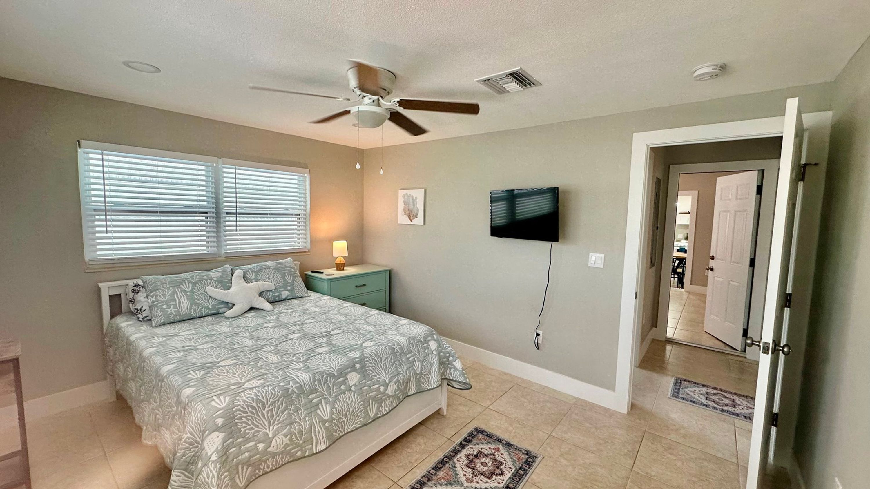 A bedroom with a bed, a ceiling fan, a TV, and a door leading to another room.