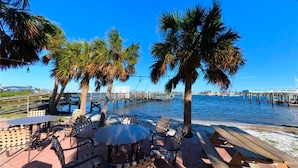 A waterfront patio with tables and chairs, palm trees, and a pier extending into the water. - Seagrape Bay 205 in Fort Myers Beach (Fort Myers Beach)