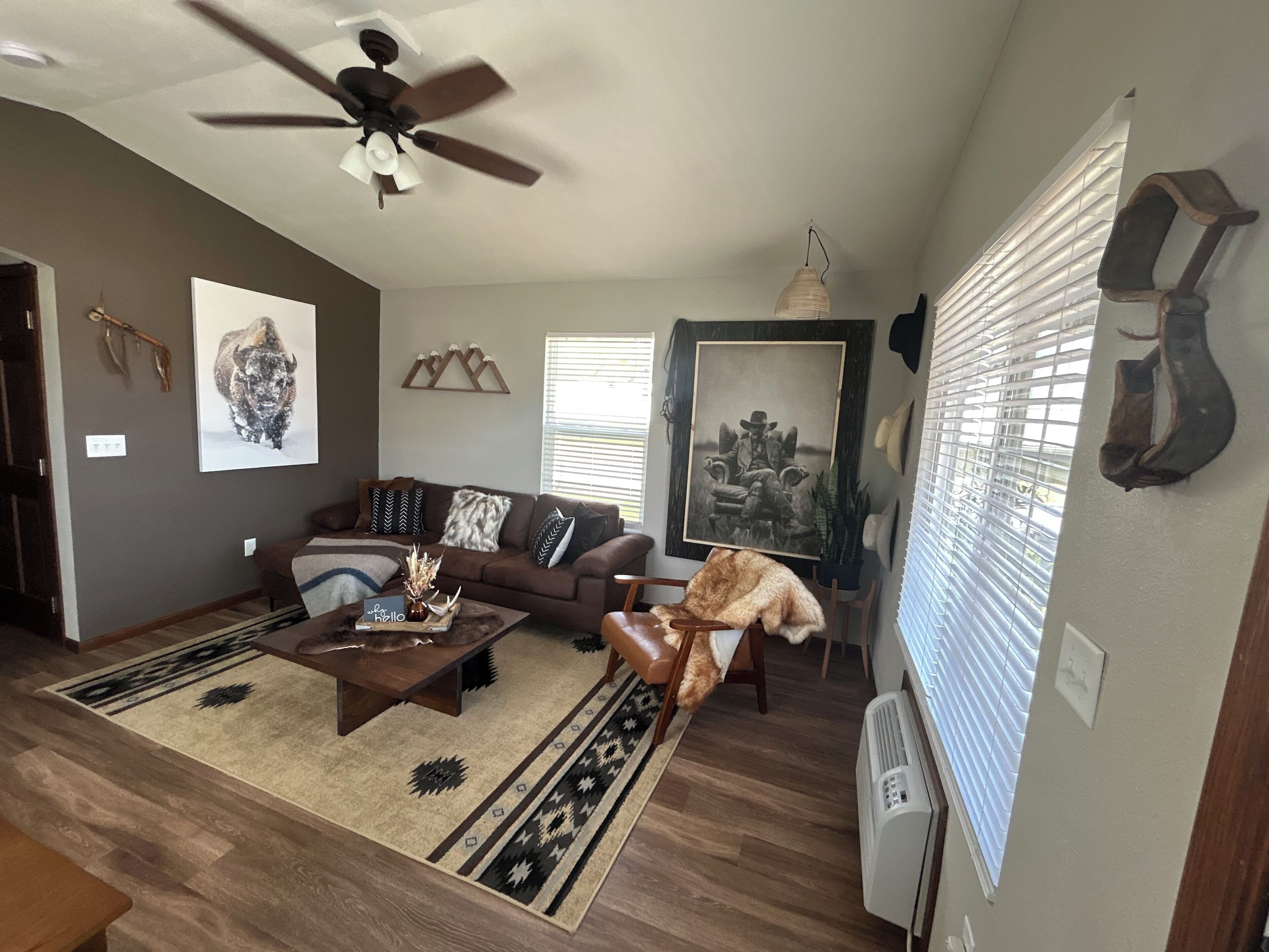 A living room with a brown sofa, a coffee table, and a ceiling fan. There are wall decorations and a window with blinds.