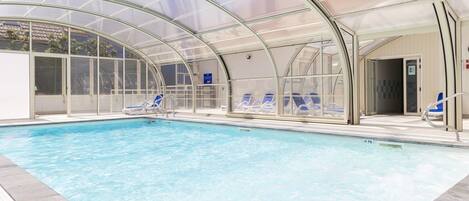 An indoor swimming pool with clear water, surrounded by blue lounge chairs and a glass-enclosed roof.