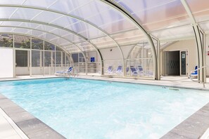 An indoor swimming pool with clear water, surrounded by blue lounge chairs and a glass-enclosed roof. - Residence Les Villas De La Baie (Le Crotoy)