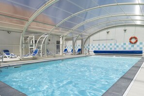 An indoor swimming pool with clear water, blue lounge chairs, a lifebuoy, and a glass ceiling structure. - Residence Les Villas De La Baie (Le Crotoy)