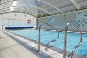 An indoor swimming pool with clear water, metal pool ladders, a blue checkered wall, and a glass ceiling. - Residence Les Villas De La Baie (Le Crotoy)