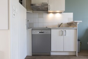 Cocina compacta con lavavajillas, armarios blancos y una campana extractora de acero inoxidable.