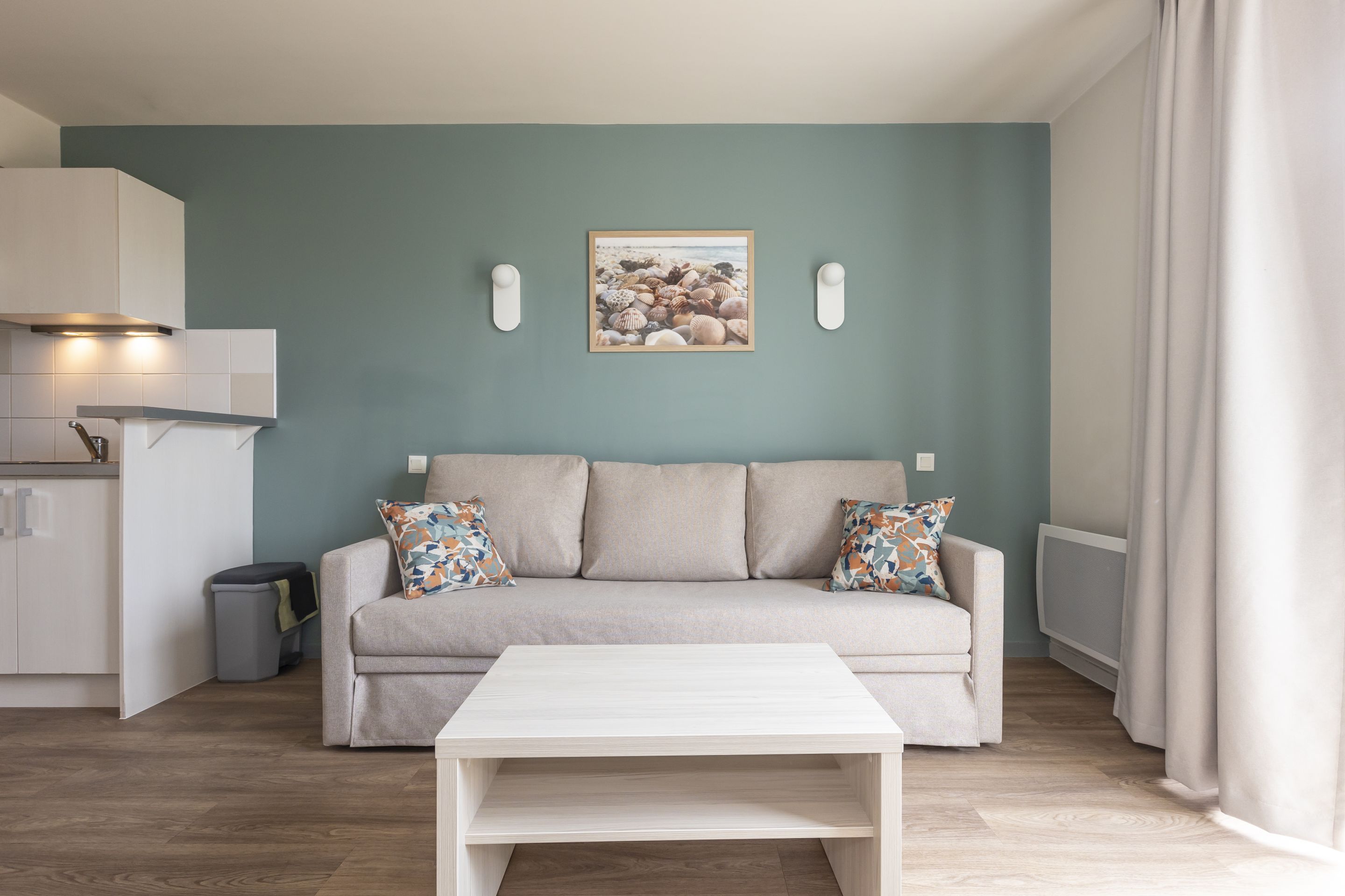 A modern living room with a beige sofa, a white coffee table, and a framed picture of seashells on the wall.