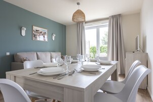 Un comedor con una mesa de madera clara preparada para cuatro, sillas blancas, una lámpara colgante de fibras naturales y vistas al exterior con follaje.