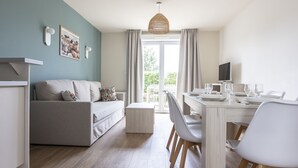A modern living room with a sofa, dining table, and chairs. There is a television on a stand and a window with curtains. - Residence Les Villas De La Baie (Le Crotoy)