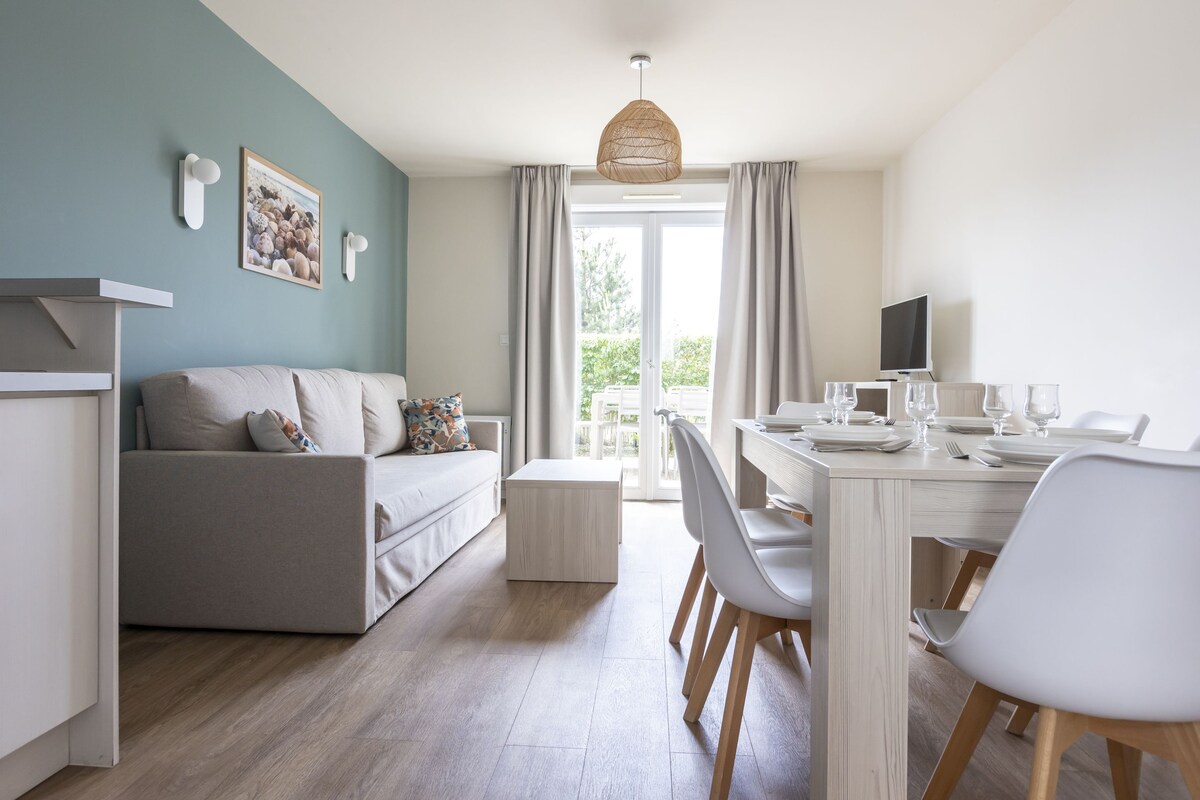 A modern living room with a sofa, dining table, and chairs. There is a television on a stand and a window with curtains.
