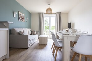 Ein modernes Wohnzimmer mit einer Couch, einem Esstisch und Stühlen. Ein Fernseher steht auf einem Schrank und ein Fenster ist mit Vorhängen drapiert.