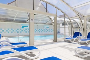 A covered swimming pool area with blue lounge chairs and a glass ceiling. - Residence Les Villas De La Baie (Le Crotoy)